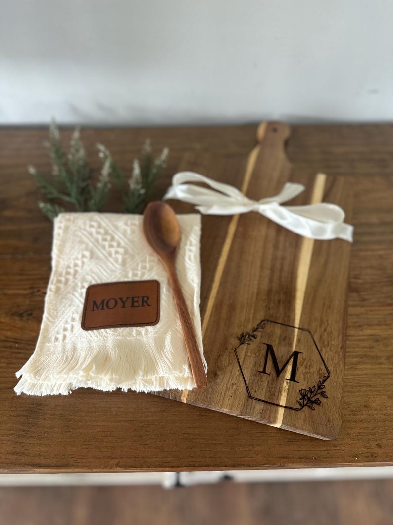 May include: A wooden cutting board with a handle, a white ribbon, and a monogrammed design. A white fringed cloth with a leather patch that says "MOYER" and a wooden spoon are next to the board. A sprig of greenery is also present.