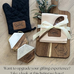 May include: A black oven mitt, wooden cutting board, coasters, and a kitchen towel with a wooden spoon. The items are labeled with "Home Sweet Home". The cutting board has a sprig of greenery and the towel is tied with a ribbon.
