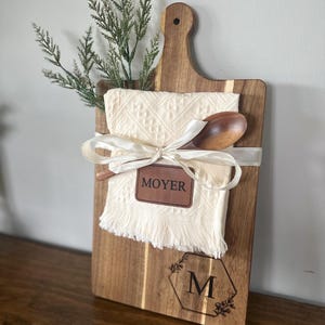 May include: A wooden cutting board with a handle, featuring a cream-colored cloth, a wooden spoon, and a decorative ribbon. The board has the name "MOYER" and the letter "M" engraved on it, along with floral accents. Greenery is placed at the top.