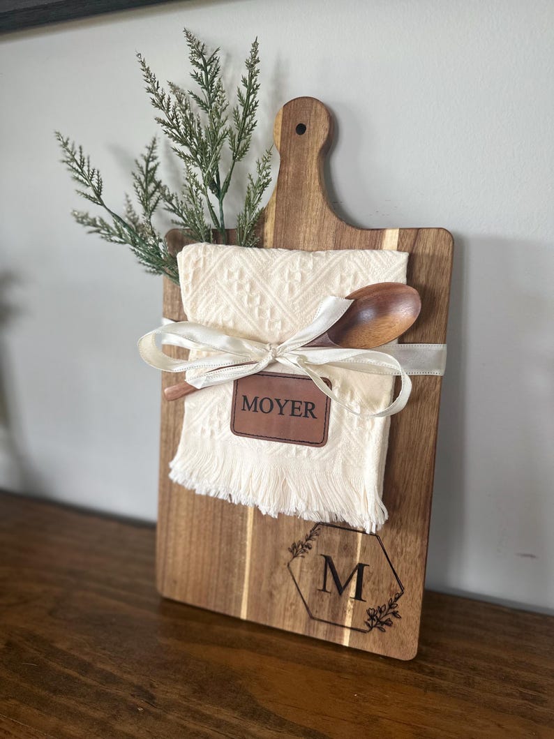 May include: A wooden cutting board with a handle, featuring a cream-colored cloth, a wooden spoon, and a decorative ribbon. The board has the name "MOYER" and the letter "M" engraved on it, along with floral accents. Greenery is placed at the top.