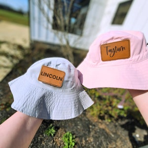 May include: Two bucket hats, one light gray and one pink, are displayed. Each hat has a brown leather patch with a name on it. The gray hat says "LINCOLN" and the pink hat says "Taytum". The hats are held up in front of a blurred outdoor background.