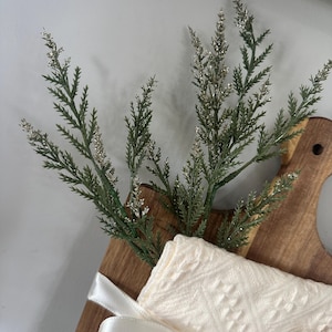 May include: A wooden cutting board with a cream-colored cloth and a sprig of green and silver decorative foliage. The cloth is tied with a white satin ribbon. The cutting board has a handle.