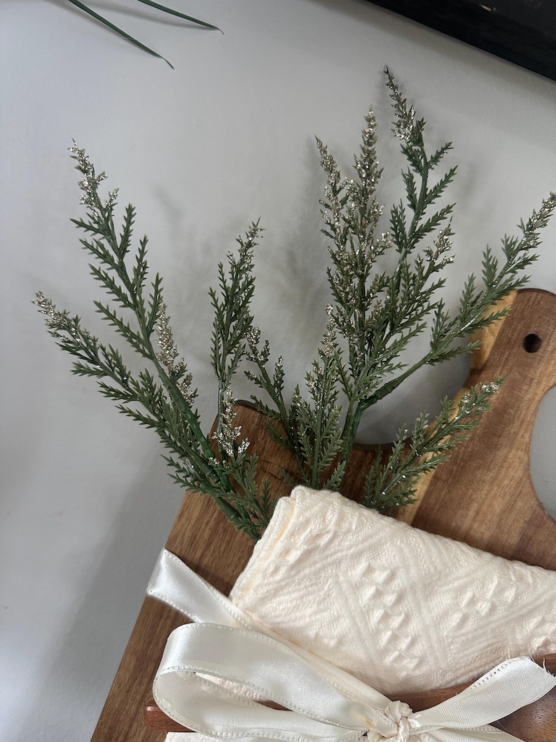 May include: A wooden cutting board with a cream-colored cloth and a sprig of green and silver decorative foliage. The cloth is tied with a white satin ribbon. The cutting board has a handle.