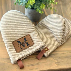 May include: Two beige and white striped oven mitts with brown leather accents. One mitt features a brown leather patch with the name "MOYER" and Roman numerals. Each mitt has a brown leather loop for hanging.