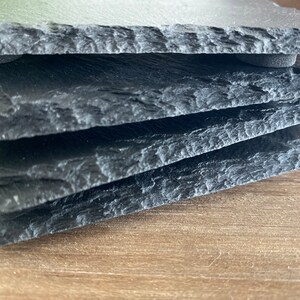 May include: A stack of four black slate coasters with rough, textured edges. The coasters are rectangular and appear to be made of natural stone. The image is a close-up, highlighting the layered texture and dark color of the slate.
