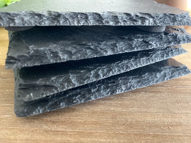 May include: A stack of four black slate coasters with rough, textured edges. The coasters are rectangular and appear to be made of natural stone. The image is a close-up, highlighting the layered texture and dark color of the slate.