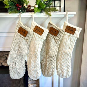 Puede incluir: Cuatro calcetines de Navidad de punto blanco con etiquetas de cuero marrón. Los calcetines están colgados en una repisa de chimenea blanca con una guirnalda verde encima. Los nombres en las etiquetas son "Daddy", "Mama", "Austin" y "Mallory".