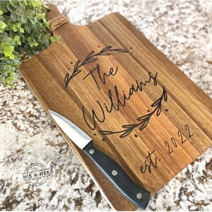 May include: Wooden cutting board with a handle wrapped in twine. The board is engraved with "The Williams" inside a leaf wreath and "est. 2022". A small knife with a black handle rests on the board.