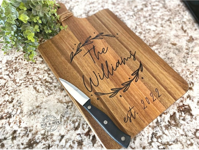May include: Wooden cutting board with a handle wrapped in twine. The board is engraved with "The Williams" inside a leaf wreath and "est. 2022". A small knife with a black handle rests on the board.