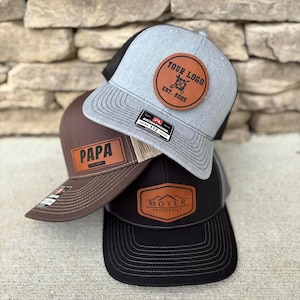 Könnte beinhalten: Drei Trucker-Caps mit braunen Leder-Patches. Die oberste Kappe ist grau mit einem schwarzen Netzrücken und einem braunen Leder-Patch mit dem Text "YOUR LOGO EST. 2025". Die mittlere Kappe ist braun mit einem schwarzen Netzrücken und einem braunen Leder-Patch mit dem Text "PAPA EST. 2019". Die unterste Kappe ist schwarz mit einem grauen Netzrücken und einem braunen Leder-Patch mit dem Text "MOYER PROPERTIES".