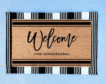 May include: A brown coir doormat with black and white striped edging. The doormat has the text "Welcome" in a black cursive font and "The Hendersons" in black text.
