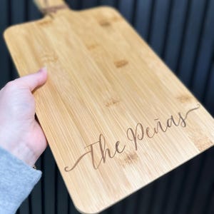 May include: A rectangular bamboo cutting board with a handle and the engraved text "The Pe&ntilde;as" in a cursive font. The board is light brown with visible wood grain and a natural finish. A jute cord is attached to the handle.