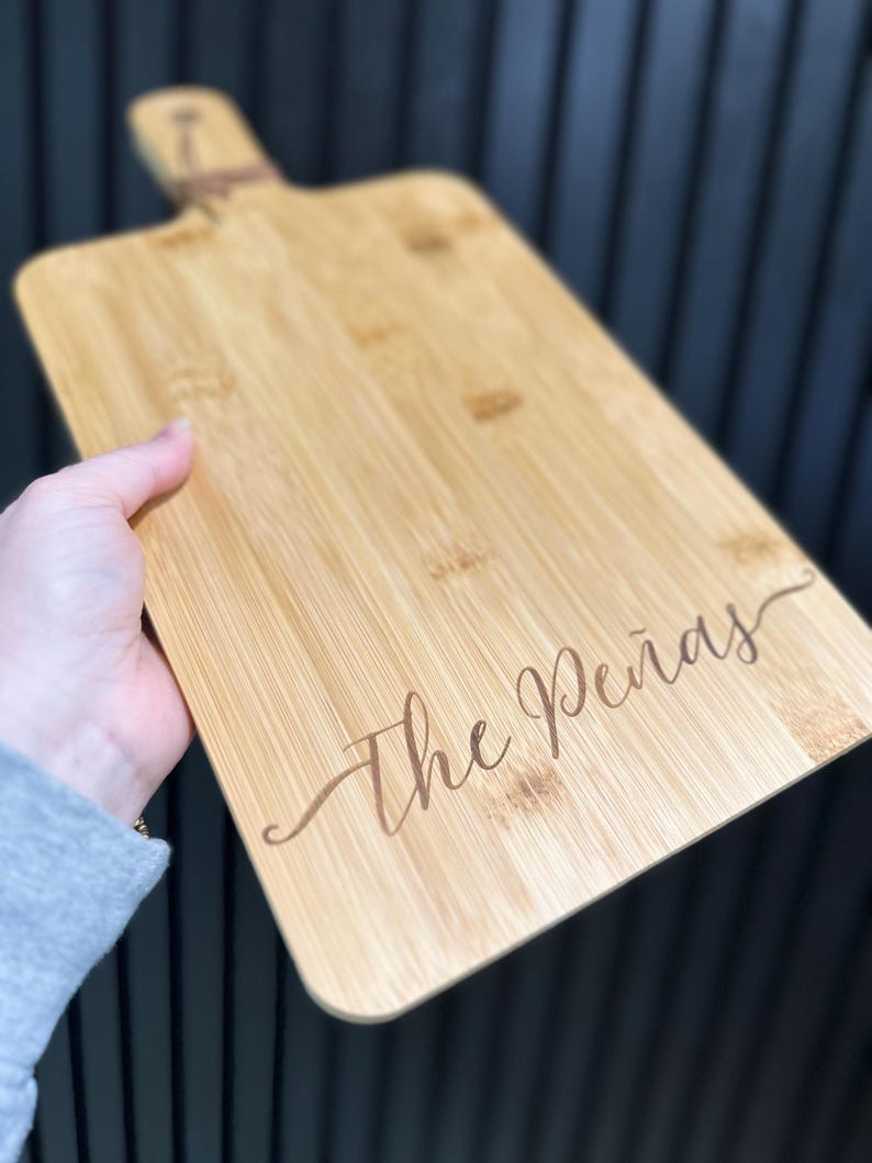 May include: A rectangular bamboo cutting board with a handle and the engraved text "The Pe&ntilde;as" in a cursive font. The board is light brown with visible wood grain and a natural finish. A jute cord is attached to the handle.