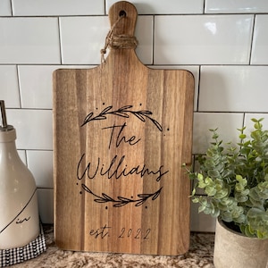 May include: Wooden cutting board with a handle and the engraved text "The Williams" inside a wreath, with "est. 2022" below. The board is light brown and sits on a countertop with white tile in the background.