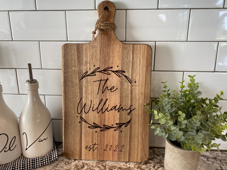 May include: Wooden cutting board with a handle and the engraved text "The Williams" inside a wreath, with "est. 2022" below. The board is light brown and sits on a countertop with white tile in the background.