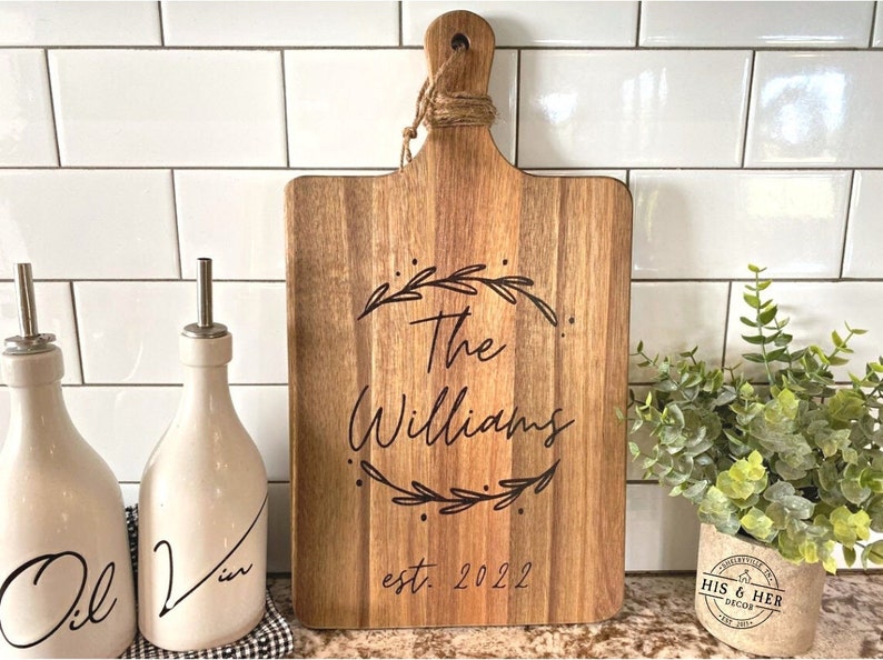 May include: Wooden cutting board with a handle, featuring the engraved text "The Williams" inside a laurel wreath, and "est. 2022" below. The board is light brown and has a natural wood grain. A decorative kitchen item.