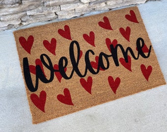May include: A brown coir doormat with the word "welcome" in black cursive lettering. The doormat is decorated with red hearts.