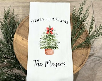 May include: White Christmas-themed kitchen towel with a watercolor Christmas tree design. The towel has the text "MERRY CHRISTMAS" above the tree and "The Moyers" below it. A red bow adorns the top of the tree.