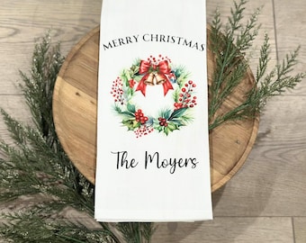 May include: White Christmas-themed kitchen towel with the text "MERRY CHRISTMAS" and "The Moyers". The towel features a wreath design with red berries, green leaves, and a red bow with golden bells. The towel is on a wooden tray.