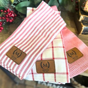 May include: Three red and white patterned kitchen towels. One towel has red stripes, another has a red and white plaid pattern, and the third has a textured red pattern. Each towel has a brown leather patch with the letter "M" and the text "MOYER EST. 2011".