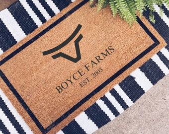 Puede incluir: Una alfombra rectangular con el texto "BOYCE FARMS EST. 2003" en negro. La alfombra tiene un centro de fibra de coco marrón con un borde negro y está colocada sobre una alfombra a rayas blancas y negras. Un helecho es visible en la esquina superior derecha.
