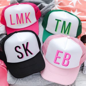 May include: Four trucker hats in pink, green, black, and light pink. Each hat has a white front panel with initials in a contrasting color: LMK, TM, SK, and EB. The hats have mesh backs and are on a gray hoodie.