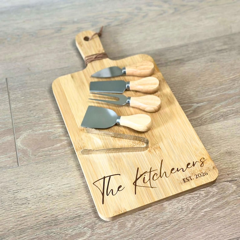 May include: A bamboo cheese board set with four stainless steel cheese tools and a pair of tongs. The board is engraved with "The Kitcheners EST. 2026" in a script font. The tools have wooden handles and are arranged on the board.