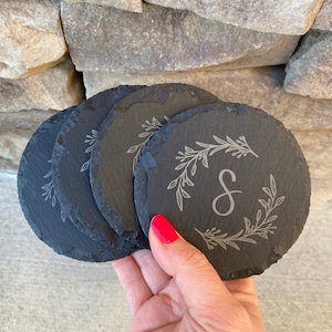 May include: A set of five round, dark gray slate coasters. Each coaster features a delicate, engraved floral wreath design with the letter "S" in the center. The coasters are held in a hand with red nail polish.