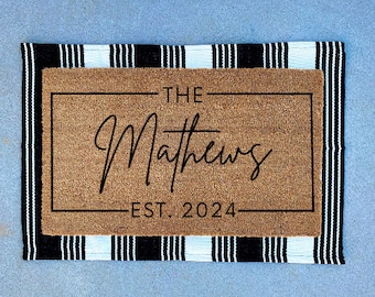 May include: A brown coir doormat with black and white striped edging. The doormat has a black frame with the text "THE Matthews EST. 2024" in black.