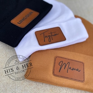 May include: Three knit beanies with personalized leather patches. The beanies are black, white, and brown. The patches have the names "Lincoln", "Taytum", and "Mama" written in cursive.