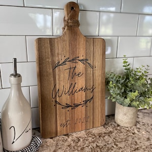 May include: Wooden cutting board with a handle and the engraved text "The Williams" inside a laurel wreath, with "est. 2022" below. The board is brown with a natural wood grain. A decorative kitchen item.