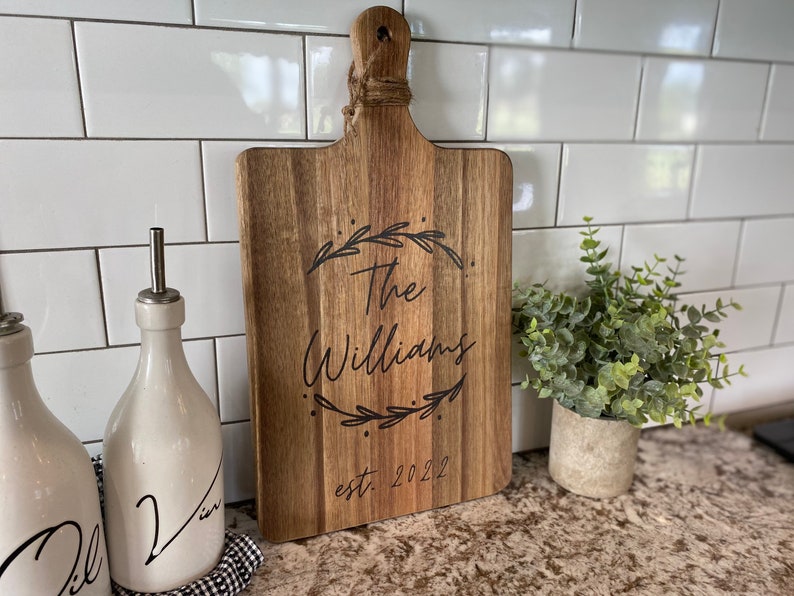 May include: Wooden cutting board with a handle and the engraved text "The Williams" inside a laurel wreath, with "est. 2022" below. The board is brown with a natural wood grain. A decorative kitchen item.