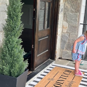 May include: A brown coir doormat with the text "The Schointuch Family" printed in black. The doormat is on a white and black striped rug in front of a wooden door.