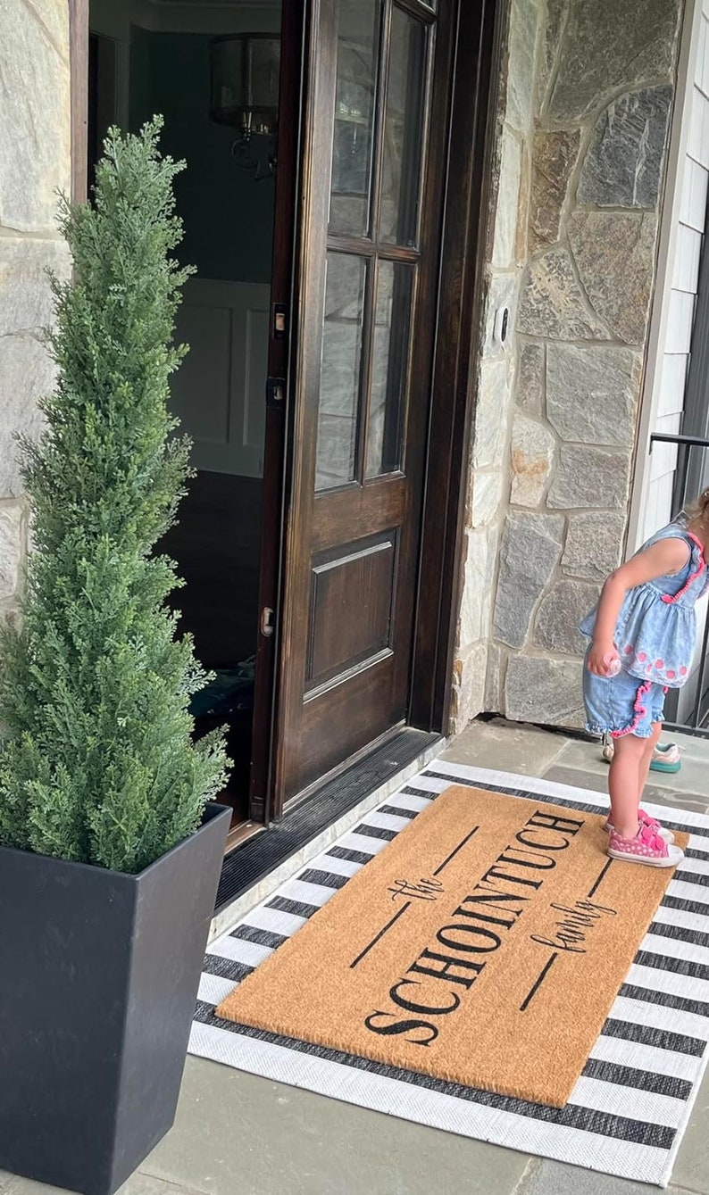 May include: A brown coir doormat with the text "The Schointuch Family" printed in black. The doormat is on a white and black striped rug in front of a wooden door.