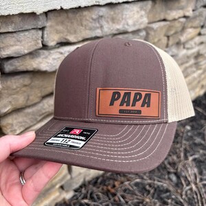 May include: Brown and beige trucker hat with a brown leather patch that says "PAPA EST. 2017".