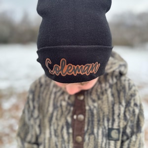 May include: Black beanie with a brown leather patch that says "Coleman" on it. The beanie is being worn by a person wearing a camouflage jacket.