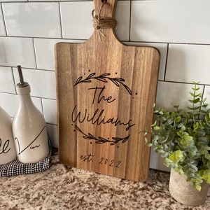May include: Wooden cutting board with a handle, engraved with "The Williams" and "est. 2022" within a leaf design. The board is made of light brown wood and has a jute rope tied through the handle.