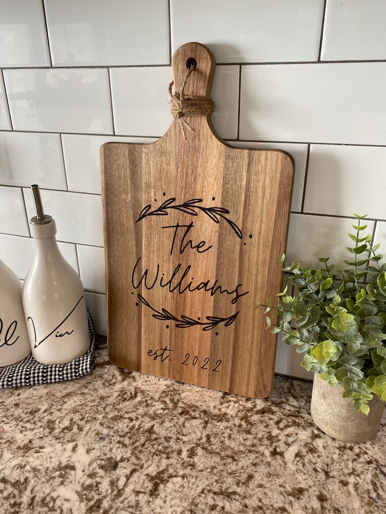 May include: Wooden cutting board with a handle, engraved with "The Williams" and "est. 2022" within a leaf design. The board is made of light brown wood and has a jute rope tied through the handle.