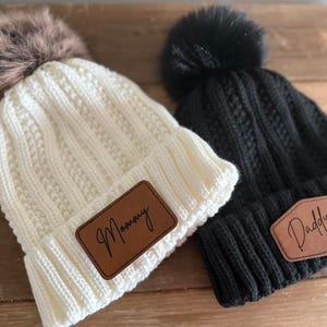 May include: Two knitted hats with faux fur pom poms. One hat is white with a brown leather patch that says "Mommy". The other hat is black with a brown leather patch that says "Daddy".