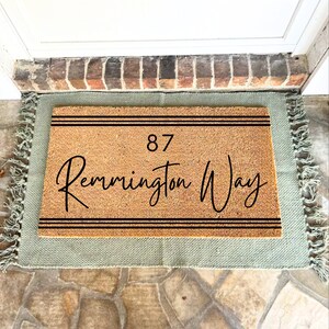 May include: A light green doormat with a brown coir insert. The insert has black text that reads "87 Remington Way" with two black lines above and below the text.