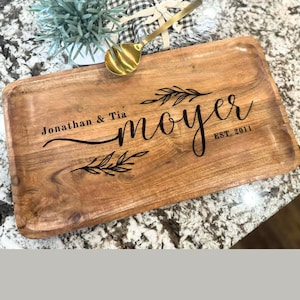 May include: A rectangular wooden serving tray with a natural wood grain finish. The tray features the names "Jonathan & Tia" and the last name "moyer" with "EST. 2011" in black script. Decorative leaf accents are also present.