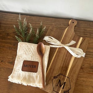May include: A wooden cutting board with a handle, tied with a white ribbon, and a wooden spoon. A white fringed towel with a leather patch that says "MOYER" is also included. The cutting board has a hexagon design with the letter "M".