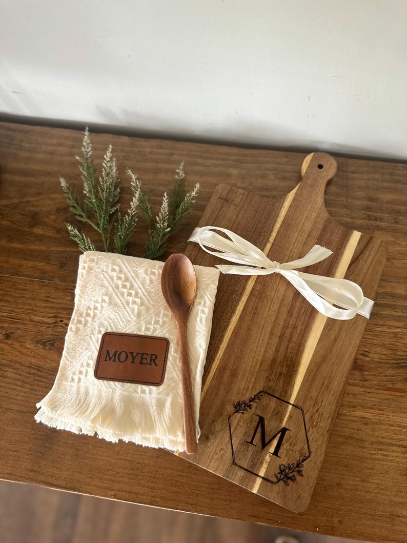 May include: A wooden cutting board with a handle, tied with a white ribbon, and a wooden spoon. A white fringed towel with a leather patch that says "MOYER" is also included. The cutting board has a hexagon design with the letter "M".