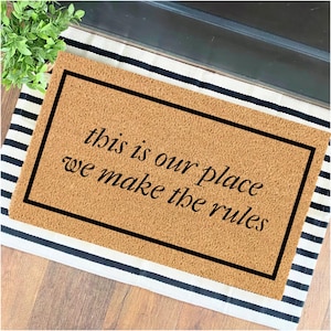 May include: A brown coir doormat with black text that reads "This is our place, we make the rules". The doormat is on a black and white striped rug.