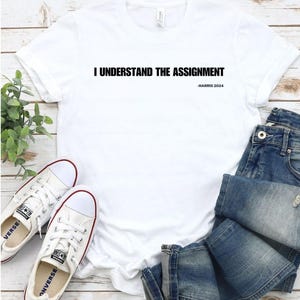 May include: White t-shirt with the text "I understand the assignment - HARRIS 2024" printed on the front.