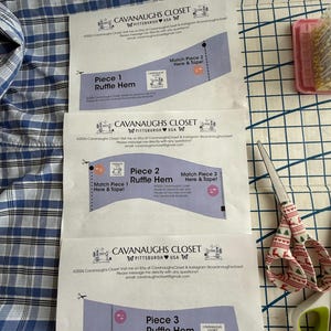 May include: Three printed paper sewing pattern sheets labeled Cavanaughs Closet for a ruffle hem, featuring instructions to match and tape pieces together. The sheets are placed on a blue and white plaid fabric next to scissors and pins.