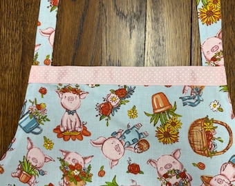 May include: Light blue apron with a repeating pattern of cartoon pigs, flowers, and gardening elements. The apron has a pink polka dot trim and straps. The pigs are wearing flower crowns and bow ties. The apron is made of fabric.