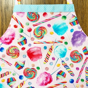 May include: Pink cotton apron with a candy theme. The apron features a variety of colorful candies, including lollipops, candy canes, and cotton candy.