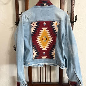 May include: Light blue denim jacket with a colorful geometric pattern on the back. The jacket has a frayed hem and cuffs with a matching patterned design. The jacket is hanging on a wooden coat rack.