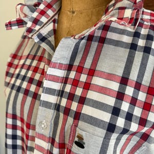 May include: A long-sleeve button-down shirt featuring a plaid pattern in red, white, black, and gray. The shirt has a ruffled collar, a front button placket, and a single chest pocket with a small embroidered stack of stones on the pocket.